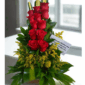 Arreglo floral vertical con rosas rojas, follaje verde, flores amarillas y tarjeta de dedicatoria sobre base rectangular