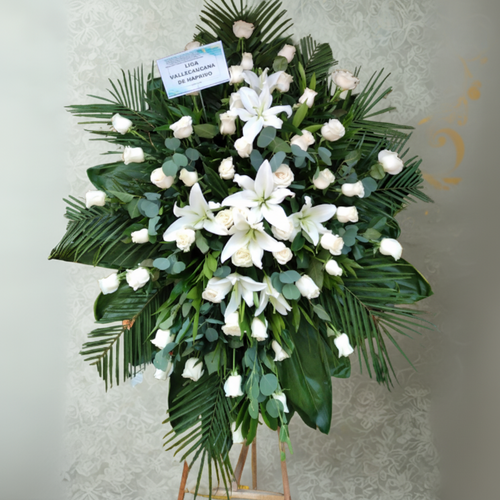 Arreglo floral fúnebre en forma de corona con lirios y rosas blancas sobre base de hojas verdes y caballete de madera
