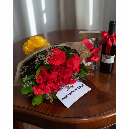 Ramo de rosas rojas envueltas en papel kraft sobre una mesa de madera, acompañado de una tarjeta blanca, una caja de bombones y una botella de vino tinto con lazo rojo