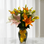 Arreglo de lirios naranjas, amarillos, blancos y rosados en un jarrón de vidrio con lazo amarillo sobre una mesa de mármol