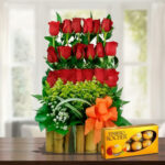 arreglo floral de rosas rojas en base de bambú con lazo naranja y caja de chocolates ferrero rocher sobre mesa