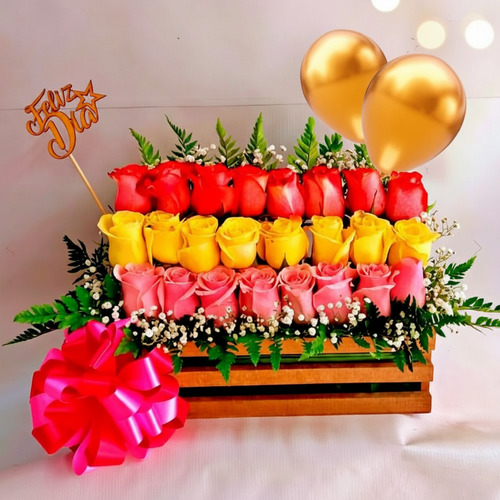 Arreglo floral en caja de madera con rosas rojas, amarillas y rosadas en tres filas, globos dorados, lazo rosa grande y letrero de Feliz Día