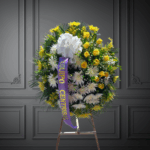 Arreglo floral en forma de corona con flores amarillas y blancas y moño blanco, montado sobre caballete de madera en fondo gris elegante