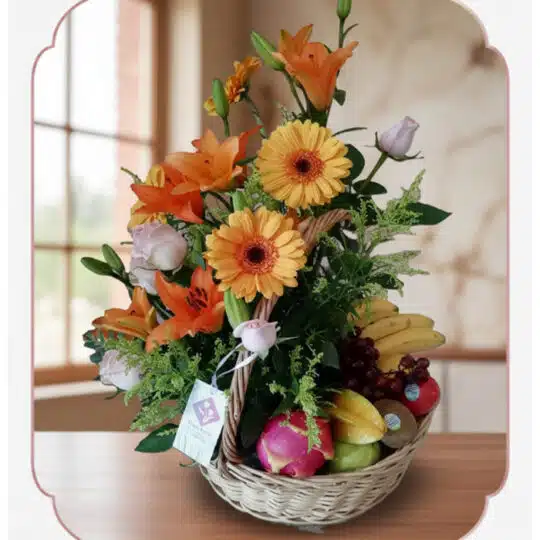 Cesta de mimbre con arreglo floral de rosas, lirios y gerberas, acompañada de frutas frescas como plátanos, uvas, manzanas y