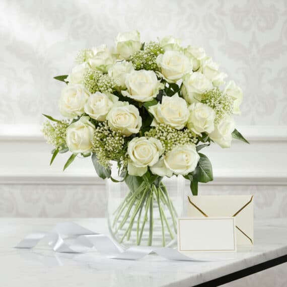 Arreglo floral Estefanía con 24 rosas blancas frescas en jarrón de cristal, tarjeta de regalo y cinta satinada sobre mármol.