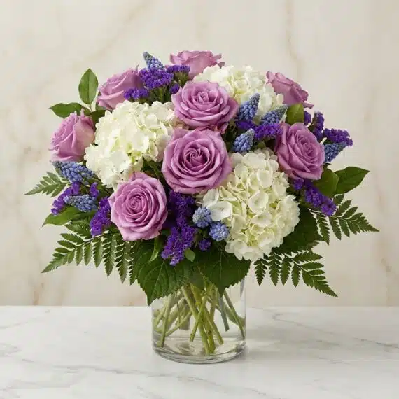 Arreglo Floral VITTORIA: Rosas Lilas y Hortensias para Regalar Arreglo floral VITTORIA con rosas lilas, hortensias blancas y muscari azul en un jarrón de cristal, sobre una mesa de mármol.