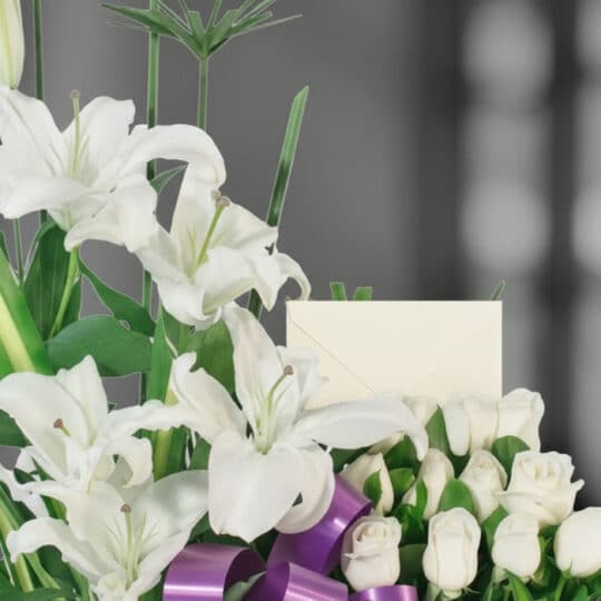 Primer plano de Arreglo Fúnebre Pureza con lirios blancos, rosas y cinta morada, rocío sutil en pétalos y follaje, profundida