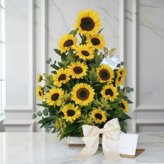 Impresionante arreglo floral con girasoles vibrantes, follaje verde y un lazo de satén, presentado sobre una mesa de mármol.