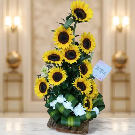 Arreglo Floral con Girasoles Sunna para Regalo Elegante Arreglo floral de girasoles Sunna en un jarrón de madera, con detalles de flores blancas y lazo verde, sobre una superficie d
