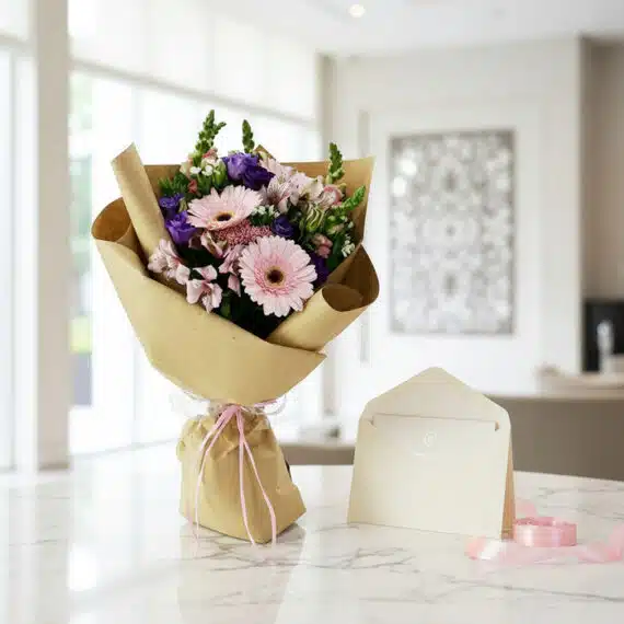 Bouquet CARINA con gerberas y astromelias rosadas, envuelto en papel kraft, sobre mesa de mármol con tarjeta de regalo y cint