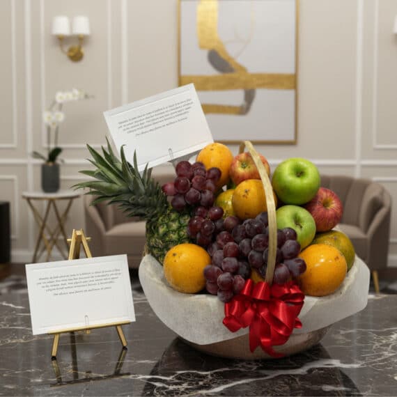 Canasta de Frutas Premium: Regalo Elegante y Saludable para Toda Ocasión Canasta de frutas premium con piña, uvas, manzanas rojas y verdes, naranjas, y un lazo rojo, sobre una mesa de mármol en un e