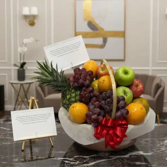 Canasta de frutas premium con piña, uvas, manzanas rojas y verdes, naranjas, y un lazo rojo, sobre una mesa de mármol en un e