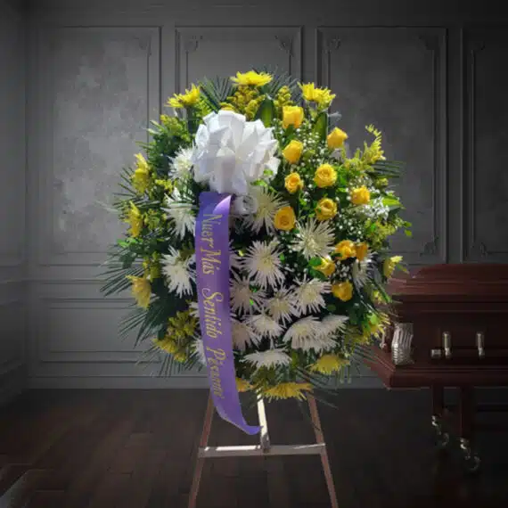 Corona fúnebre 'Estamos Contigo' con flores amarillas y blancas, cinta morada con mensaje de condolencia, junto a un ataúd en