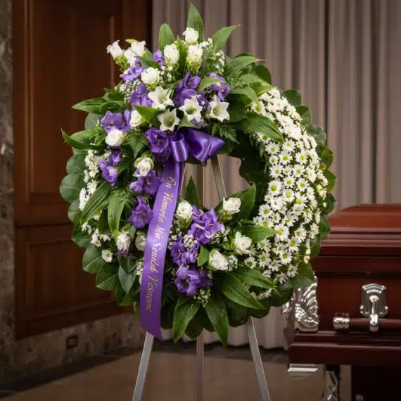 Corona fúnebre 'Homenaje a Daniel' con flores blancas y moradas, cinta morada con mensaje, en sala de velación elegante junto