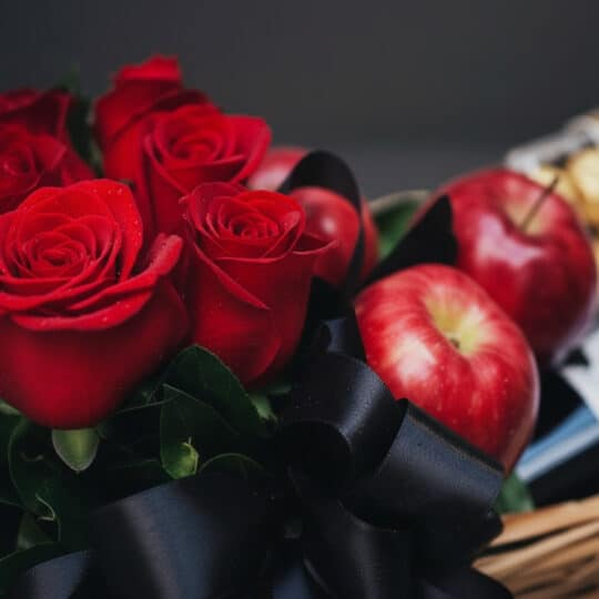 Primer plano de rosas rojas frescas con gotas de rocío, manzanas rojas brillantes y un lazo negro de satén en una cesta de re